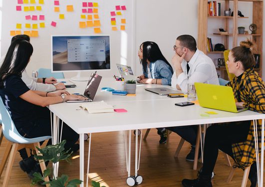 Five diverse people sit around a white table in a meeting room, focused on a large screen displaying a digital interface, with colorful sticky notes on the wall behind them.