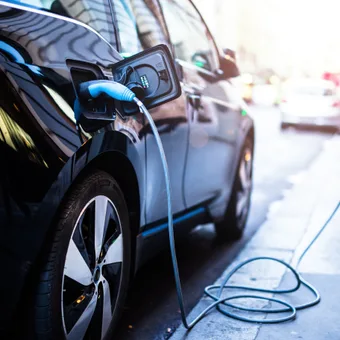 A black electric car connected to a charging station on a city street during daylight, with other vehicles and bright sunlight in the background.