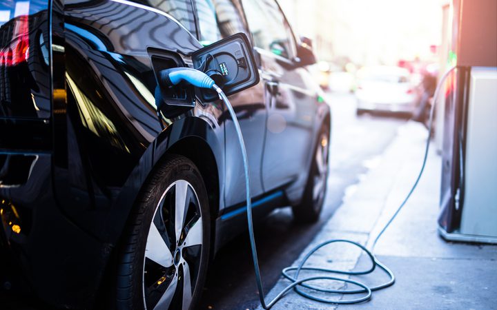 A black electric car connected to a charging station on a city street during daylight, with other vehicles and bright sunlight in the background.
