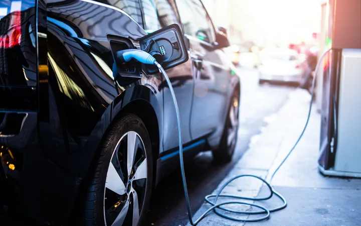 Electric car charging at a station on a city street during daytime, with other vehicles in the background and sunlight shining from the right.
