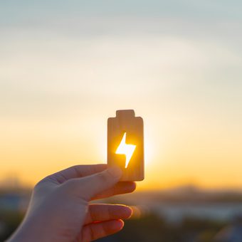 A hand holds a small wooden card with a lightning bolt cut-out, sunlight shines through it against a sunset sky.
