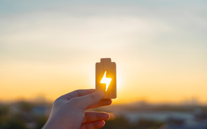 A hand holds a small wooden card with a lightning bolt cutout; warm sunlight shines through the hole as a sunset paints the sky.