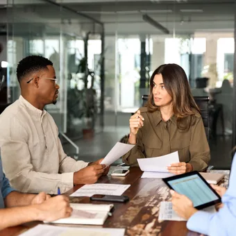 A diverse group of five people having a discussion around a table in a modern office, with women and men listening and taking notes.