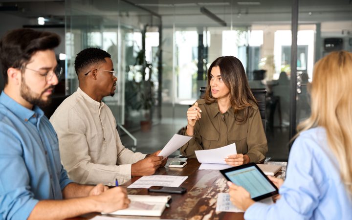 A diverse group of five people having a discussion around a table in a modern office, with women and men listening and taking notes.