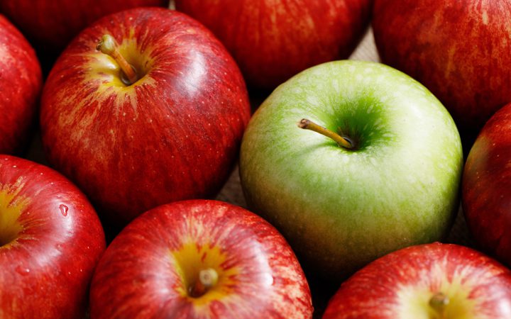 A close-up of a group of red apples with one bright green apple in the centre, all arranged on a wooden surface.