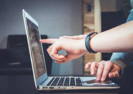 Person's hand pointing at a laptop screen, with another person's hand resting on the laptop, in a cozy indoor setting.