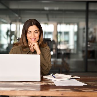 A person with shoulder-length brown hair sits at a wooden desk with a white laptop, notebook and papers in a glass-walled office.