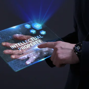 Translucent tablet showing "INTELLECTUAL PROPERTY" with blue holographic icons, held by a person in a dark suit; hexagonal pattern glow around the device.