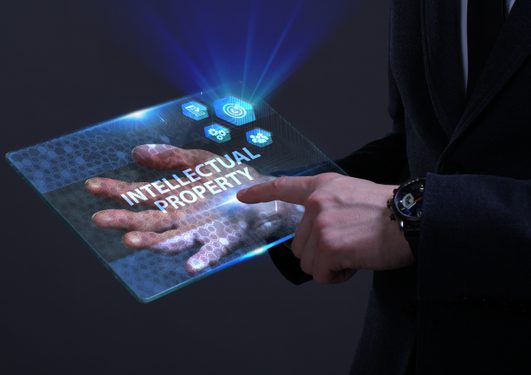 Personne en costume noir tenant une tablette transparente affichant « INTELLECTUAL PROPERTY » avec des icônes bleues holographiques et motif hexagonal.