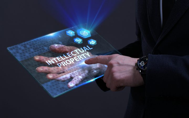 Person in a dark suit holds a transparent holographic tablet displaying "INTELLECTUAL PROPERTY" with blue icons and a hex pattern.