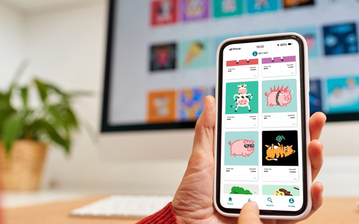 Hand holding a smartphone showing a grid of cartoon NFT artworks; blurred monitor with colourful posters and a potted plant in the background.