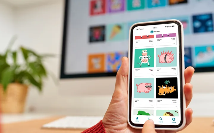 Hand holding a smartphone showing a grid of cartoon NFT artworks; blurred monitor with colourful posters and a potted plant in the background.