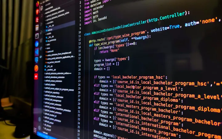 A computer screen displaying colourful Python code in a dark IDE, with a blurred background of a desk and yellow wall.
