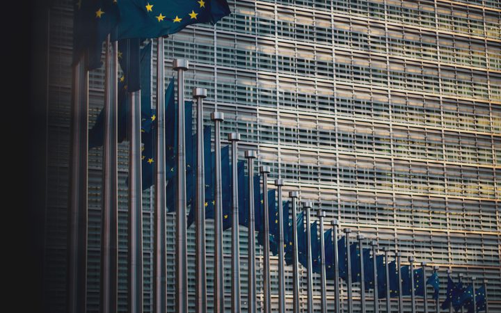 Multiple European Union flags on flagpoles in front of a modern glass building with a grid-like facade.
