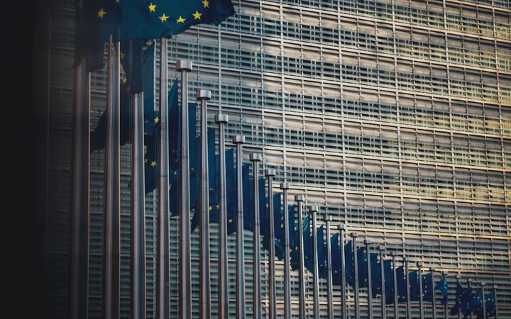 Multiple European Union flags on silver poles in front of a modern glass building facade.