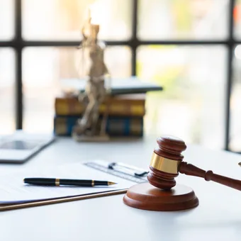 A wooden judge's gavel and brass balance scale on a white desk, with legal documents, pen, and blurred books and a skeleton figure in the background.
