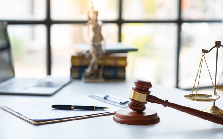 A wooden judge's gavel and brass balance scale on a white desk, with legal documents, pen, and blurred books and a skeleton figure in the background.