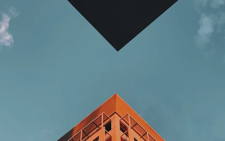 The photo shows the corner of an orange building against a blue sky with a few clouds, seen from a low angle with a black geometric shape in the background.