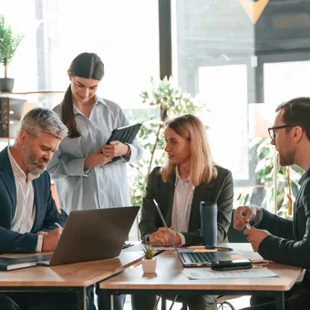 Quattro professionisti in abiti formali discutono attorno a un tavolo con laptop in un ufficio luminoso con piante e scaffali.