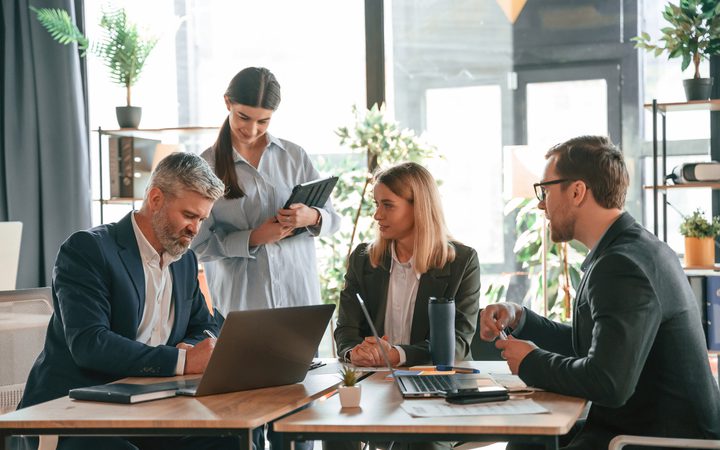 Quattro professionisti in abiti formali discutono attorno a un tavolo con laptop in un ufficio luminoso con piante e scaffali.