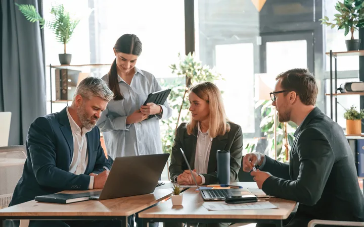 Quattro professionisti in abiti formali discutono attorno a un tavolo con laptop in un ufficio luminoso con piante e scaffali.