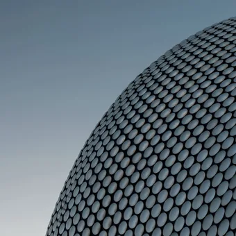 A modern building with a curved façade covered in numerous evenly spaced, silver circular tiles against a clear sky.