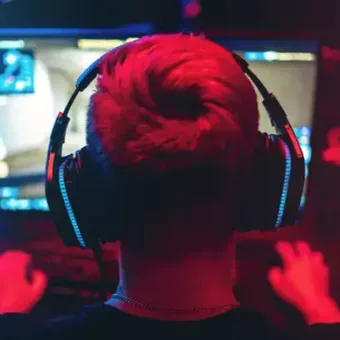 Person with bright hair wearing headphones, playing video games at a screen in a dark room with red and blue lighting.