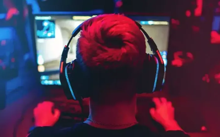 Person with bright hair wearing headphones, playing video games at a screen in a dark room with red and blue lighting.