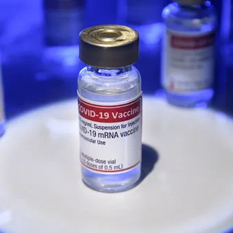 A vial of COVID-19 vaccine with a red and white label, surrounded by other vials under blue lighting, on a white circular surface.