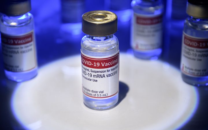 A vial of COVID-19 vaccine with a red and white label, surrounded by other vials under blue lighting, on a white circular surface.