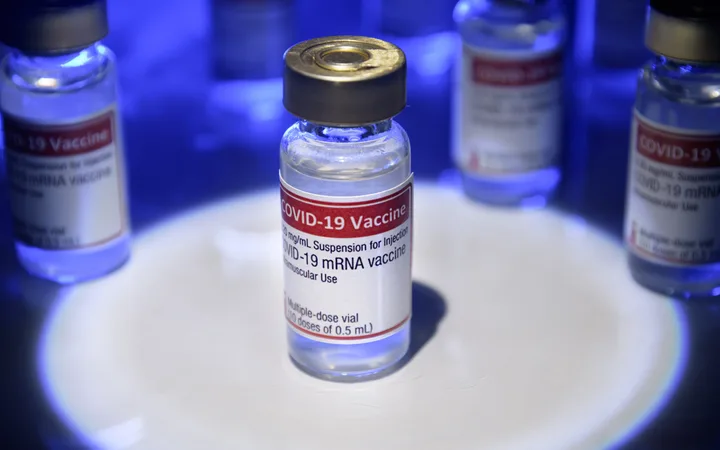 A vial of COVID-19 vaccine with a red and white label, surrounded by other vials under blue lighting, on a white circular surface.
