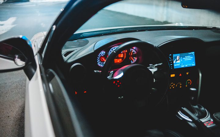View of a car's interior at night, showing illuminated dashboard, steering wheel, and infotainment screen with a parking lot in the background.