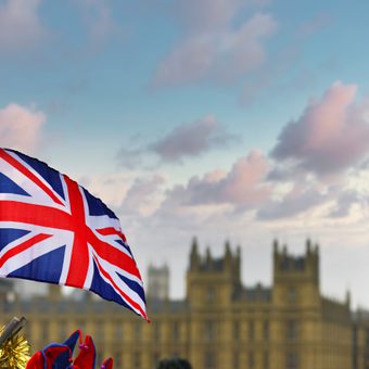 Union Jack flag waving in the foreground, with the Houses of Parliament and Big Ben blurred in the background against a blue sky.