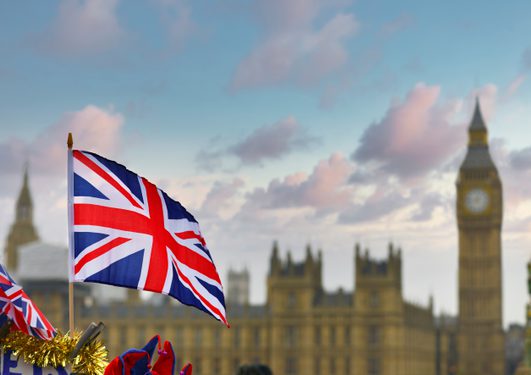 Union Jack flag waving in the foreground, with the Houses of Parliament and Big Ben blurred in the background against a blue sky.