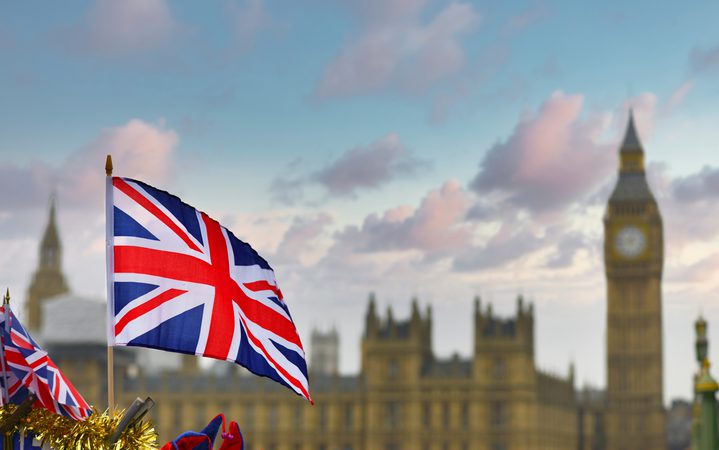 Union Jack flag waving in the foreground, with the Houses of Parliament and Big Ben blurred in the background against a blue sky.