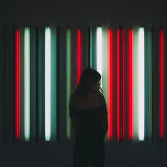 A woman with long hair stands in front of a colorful neon light installation with vertical red, teal, and white tubes in a dark gallery setting.