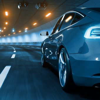 A sleek blue car driving through a lit tunnel, captured from the rear side with motion blur on the road and background lights.