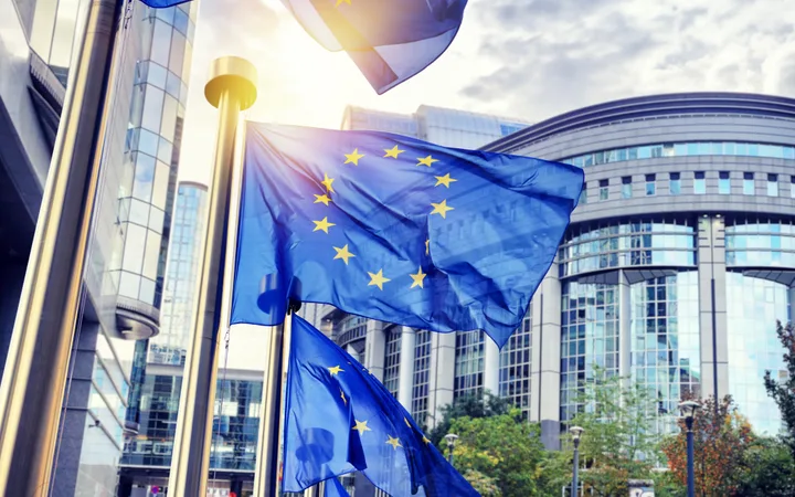 European Union flags flying outside modern glass buildings with sunlight shining through, surrounded by trees and street lamps.