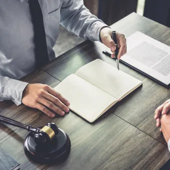 A person in a grey striped shirt and black tie taking notes in a lined notebook during a meeting, with legal documents and a gavel on a wooden table.