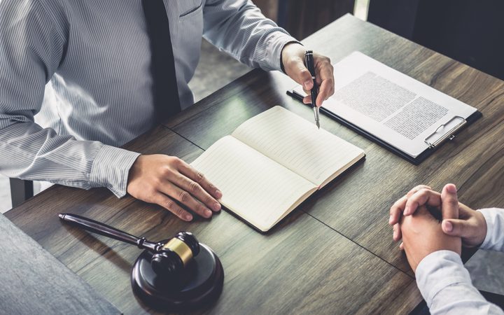 A person in a striped shirt and tie is signing a document at a wooden table, with an open notebook, a gavel, and a clipboard with a printed paper.