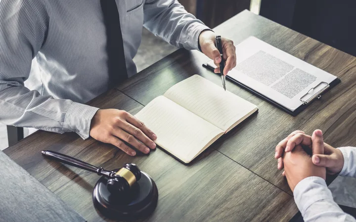 A person in a striped shirt and tie is signing a document at a wooden table, with an open notebook, a gavel, and a clipboard with a printed paper.