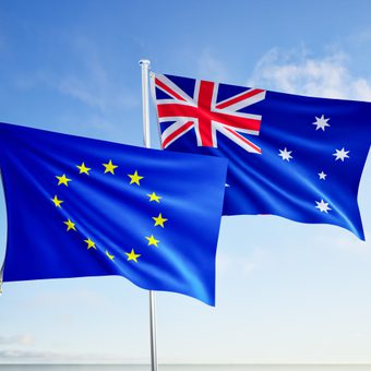 Two flags flutter in the wind: the European Union flag with a circle of yellow stars, and the Australian flag with the Union Jack, against a blue sky over the sea.