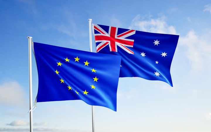 Two flags flutter in the wind: the European Union flag with a circle of yellow stars, and the Australian flag with the Union Jack, against a blue sky over the sea.