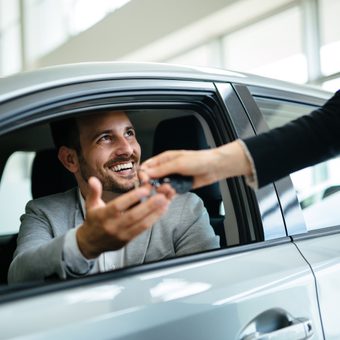A person wearing a light grey blazer sits in a car, smiling as someone hands them car keys through the open window.