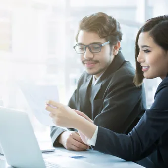 Two professionals, a man with glasses and a woman, are sitting at a table looking at documents in a bright office.