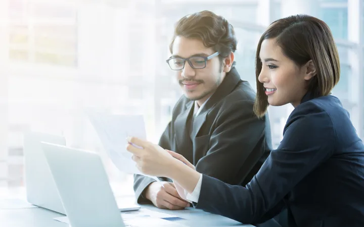 Two professionals reviewing documents together in an office, with a laptop open on the table, both smiling.