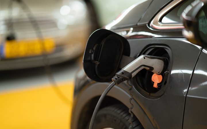 An electric car being charged with a plugged-in charging cable, with another vehicle blurred in the background at a charging station.