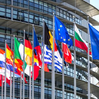 Flags of various European countries flying in front of a modern glass building with curved balconies.