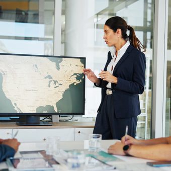 A presenter in a navy suit explains a world map on a monitor to two colleagues in a glass-walled office conference room.