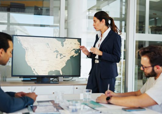 A person in a dark suit presents a world map on a monitor to two colleagues at a glass-walled office conference table.